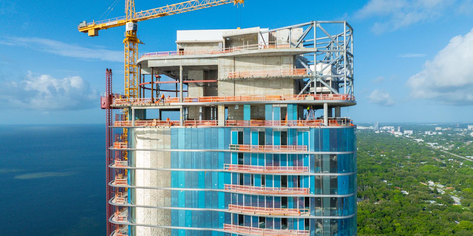 Aerial view of a tall, cylindrical high-rise under construction with blue-tinted glass windows, set near a coastal area with surrounding low-rise buildings.