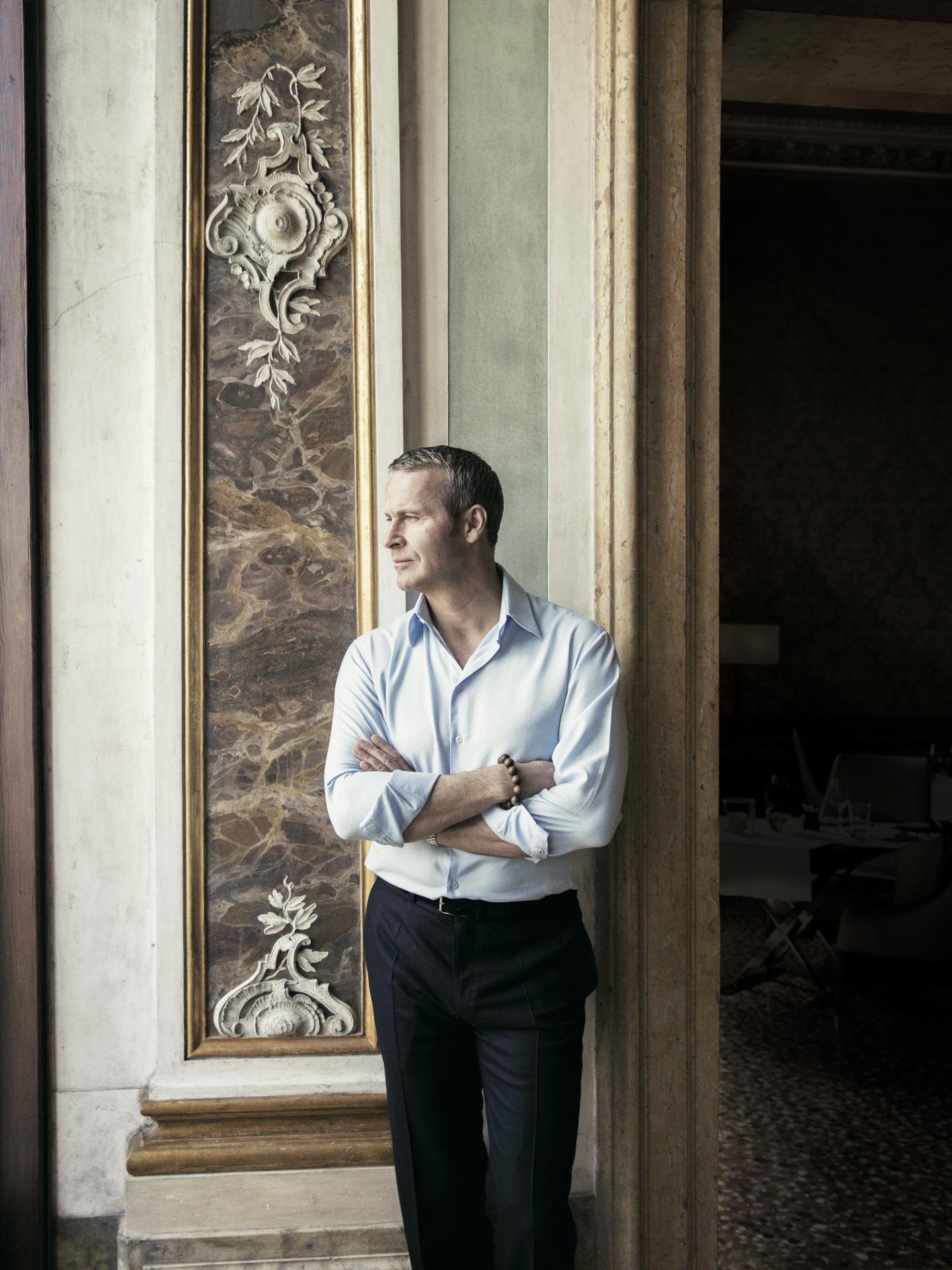 A man in a light blue shirt stands by a window with crossed arms in a historic interior.